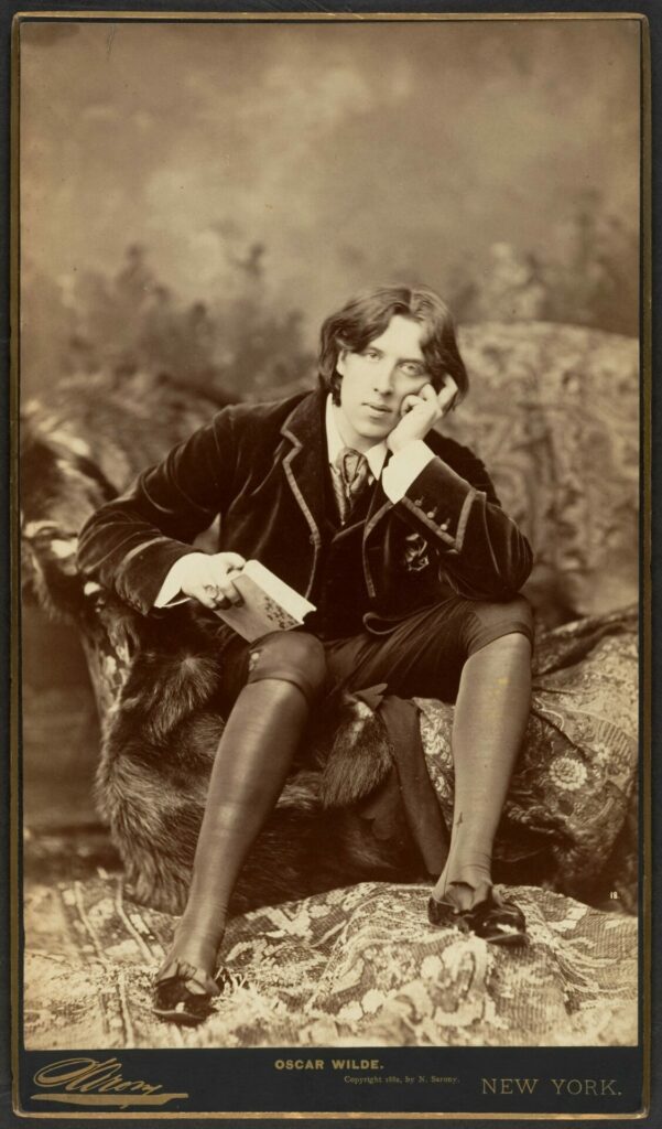 The image depicts a sepia-toned portrait of a young Oscar Wilde, seated on an ornate, patterned sofa adorned with a fur throw. He is dressed in a velvet jacket, a vest, and knee-length breeches, with stockings and formal shoes. Wilde holds a book in his left hand, resting his head on his right hand in a pensive pose, gazing directly at the viewer. The photograph exudes a sense of elegance and intellectual sophistication, characteristic of Wilde’s persona.